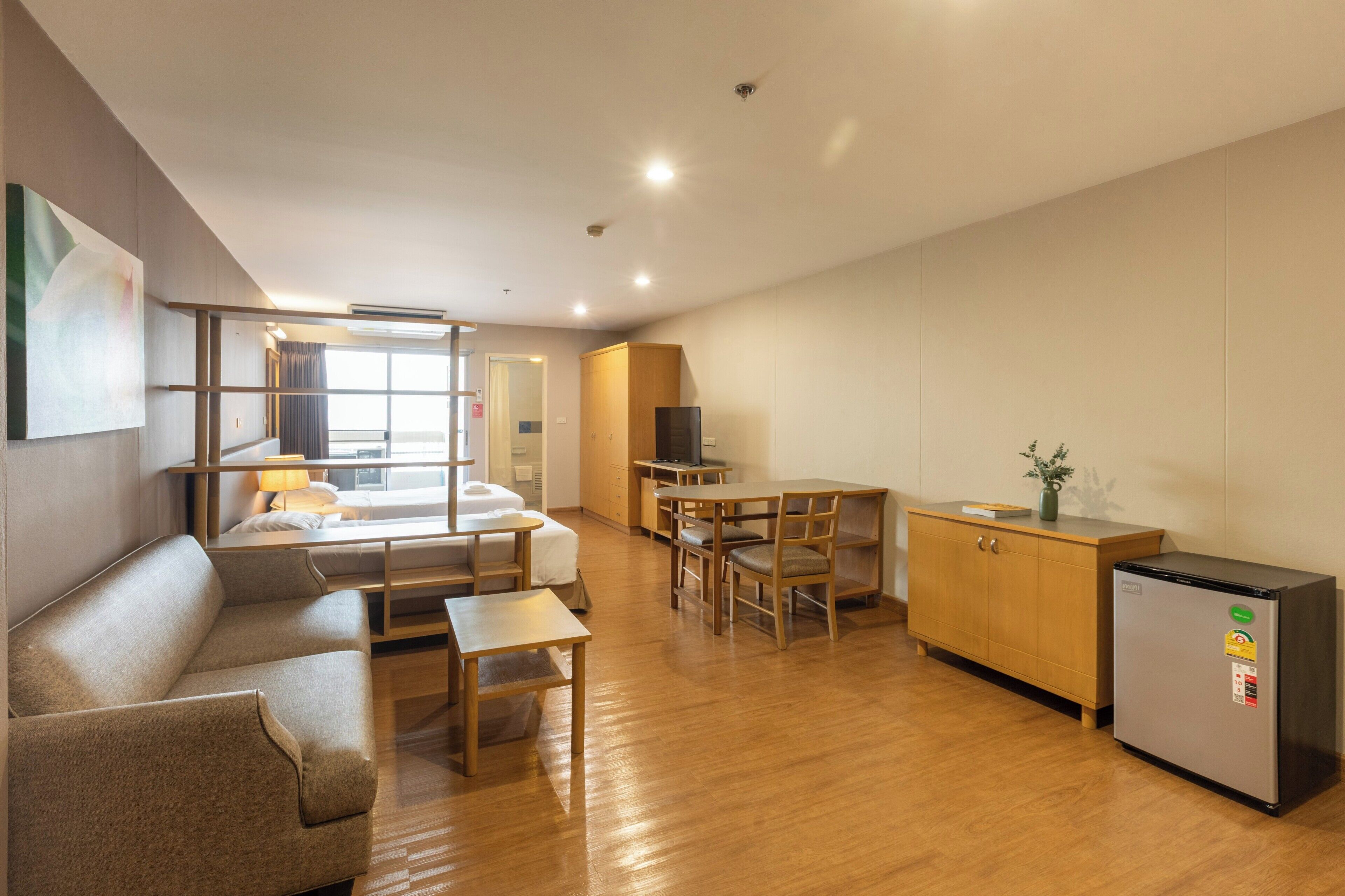A modern hotel room with a sofa, a dining table, a kitchen area, and a television.