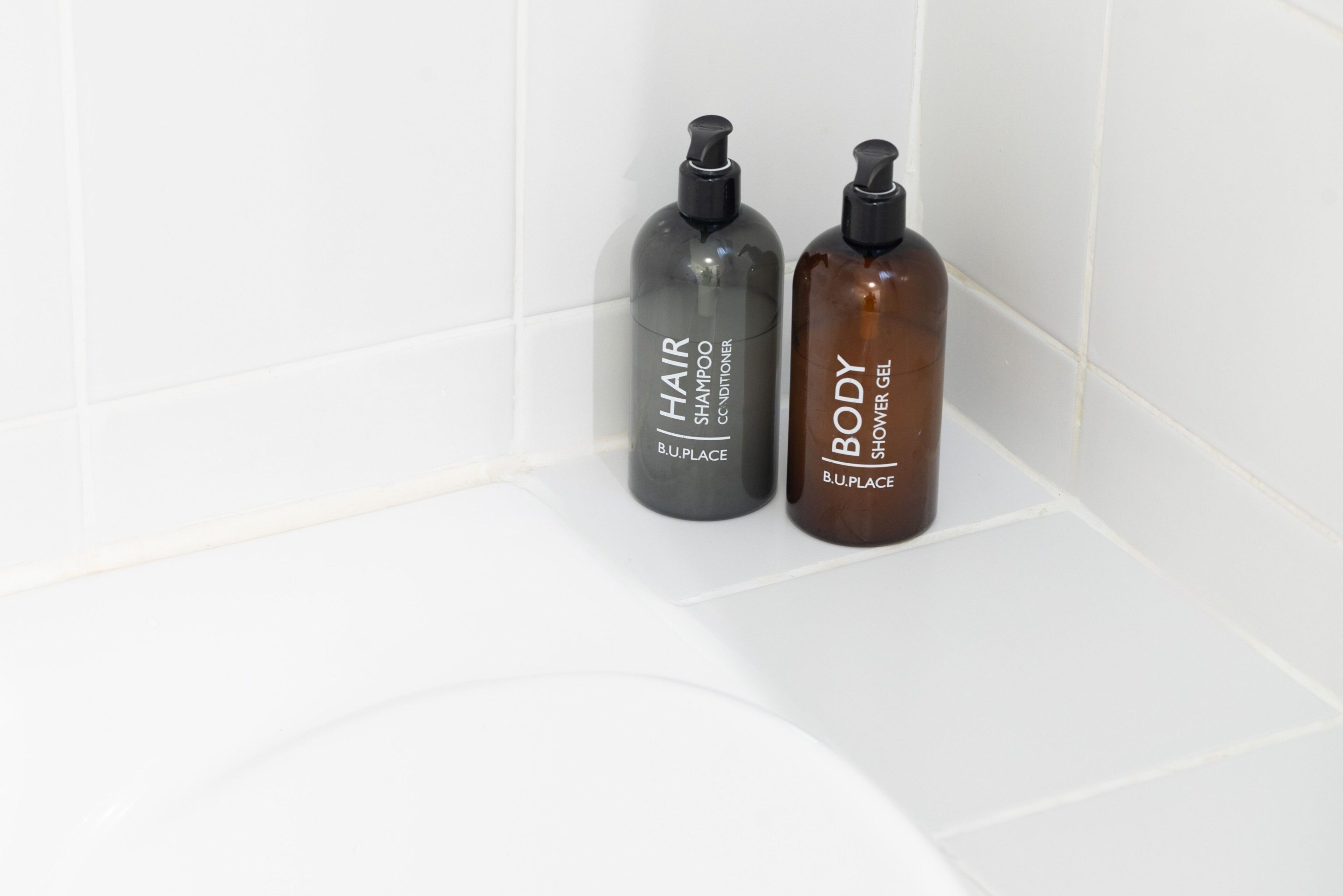Two bottles of B.U.PLACE hair and body shampoo and conditioner on a white tiled surface.