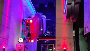 Ein neonbeleuchteter Korridor mit einem schwebenden Oldtimer, einer Zapfsäule und einer Speisekarte, die Bierpreise auflistet.