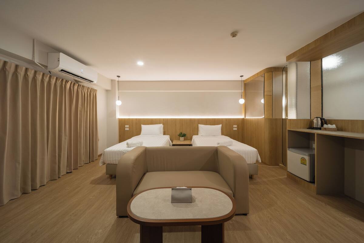 A modern hotel room with a sofa bed, a small table, a bedside table with a lamp, and a minibar.