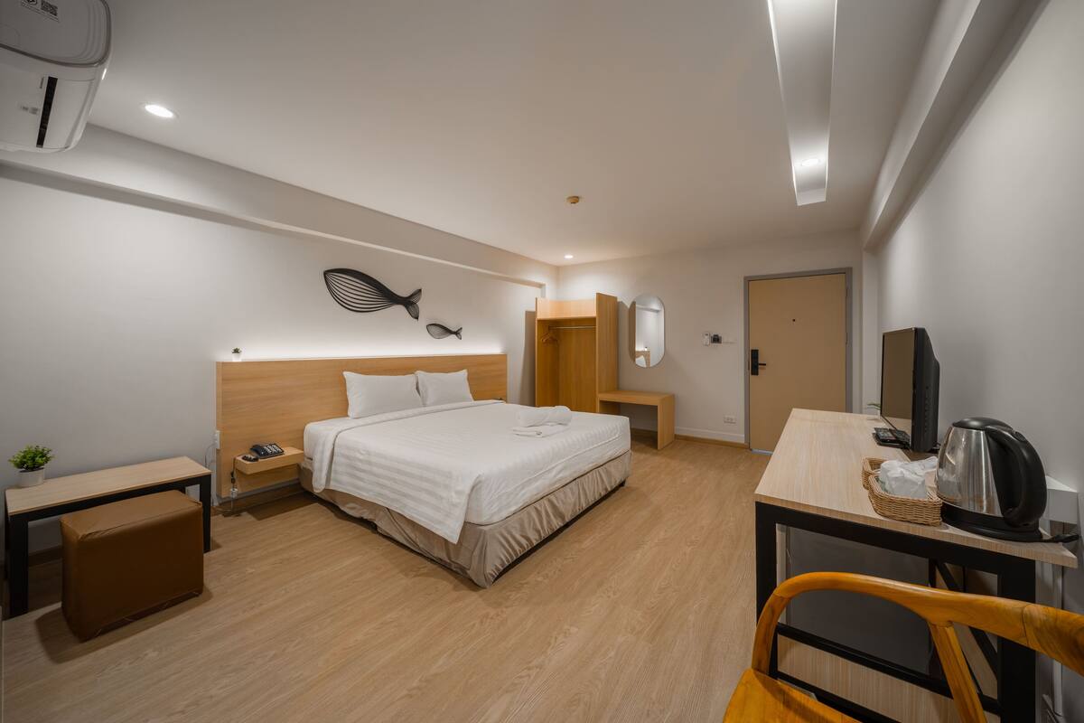 A modern hotel room with a wooden bed, bedside tables, a desk with a kettle, and a mirror.