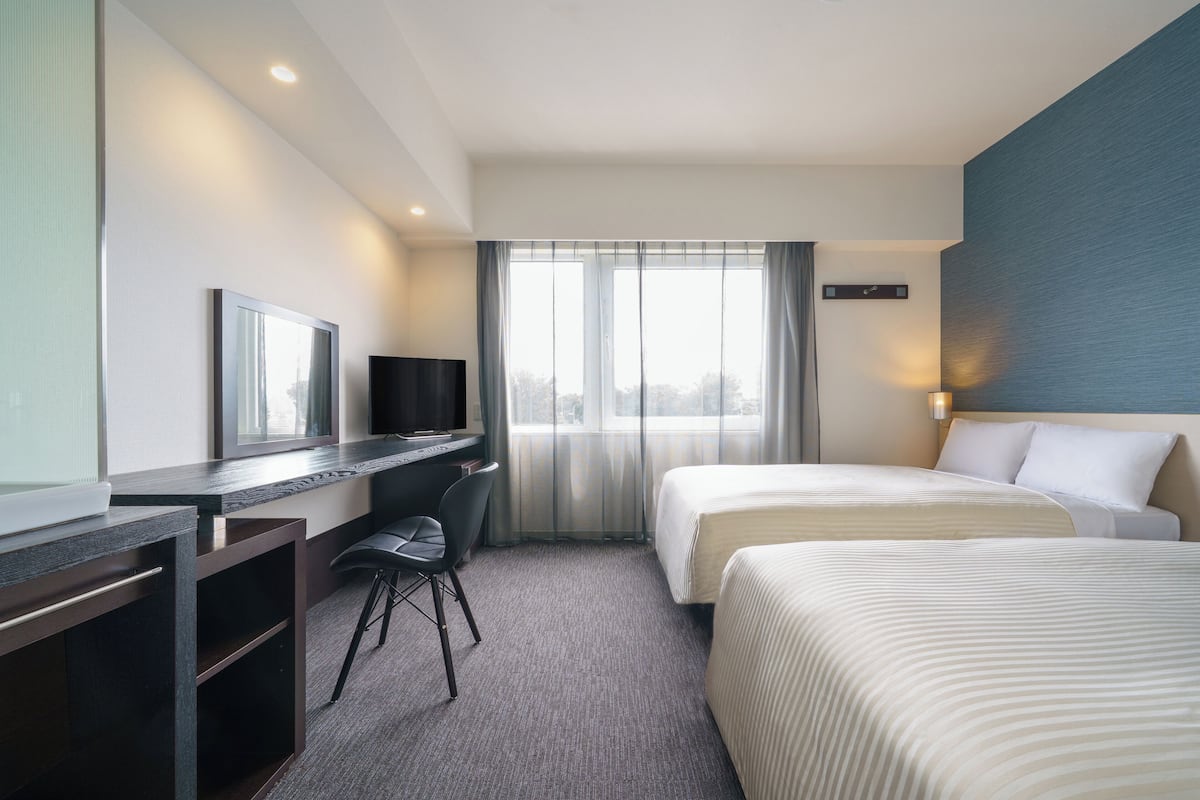 Una habitación de hotel con escritorio, dos camas, televisor y un amplio ventanal con cortinas.