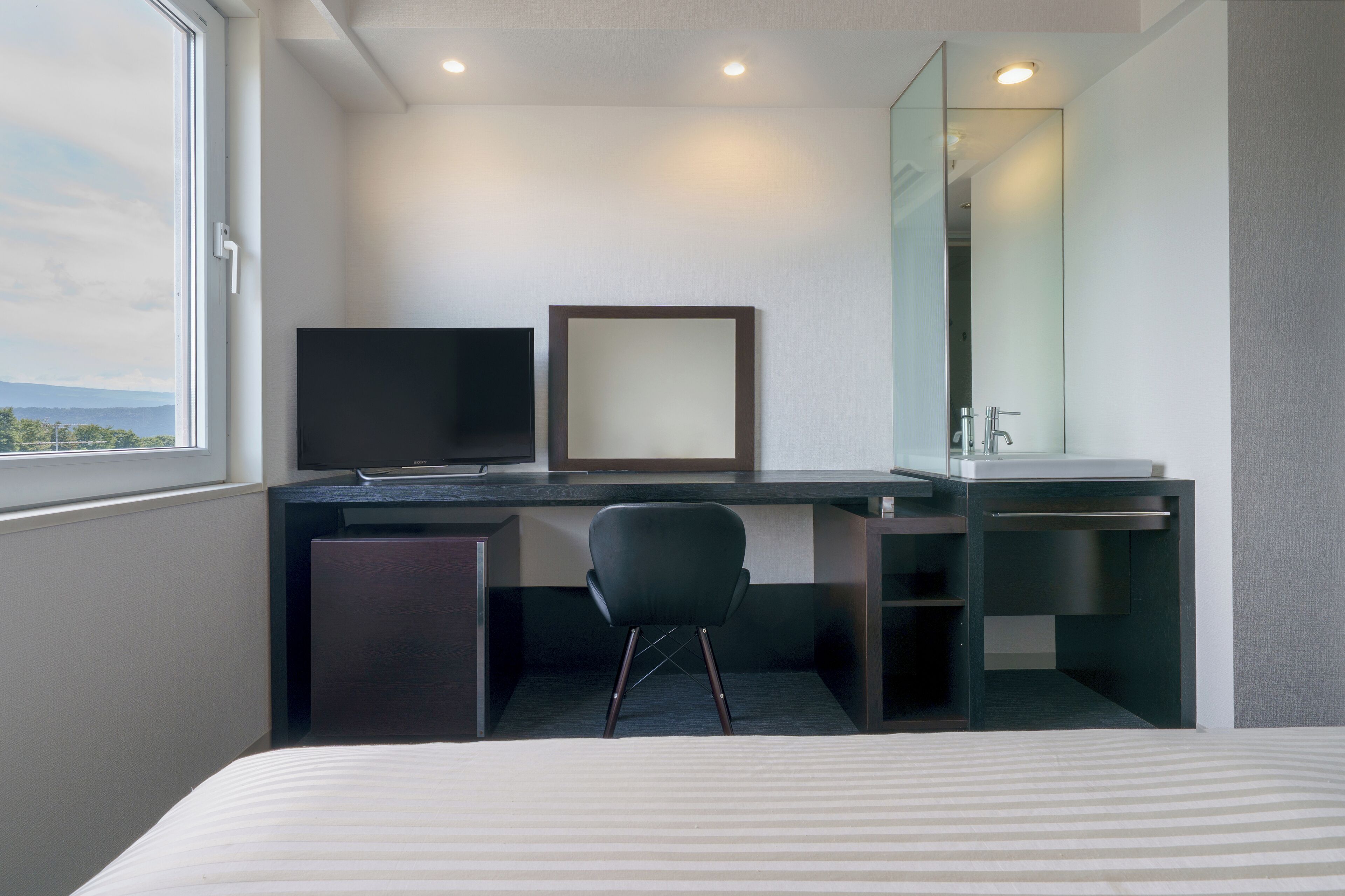 A hotel room with a bed, a desk with a chair, a television, a mirror, and a window with a view of the outdoors.