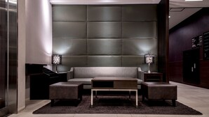 Un salon moderne avec un canapé gris, deux poufs assortis et une table basse. On y trouve deux lampes sur des tables d’appoint, un tapis foncé et une grande tête de lit rembourrée.