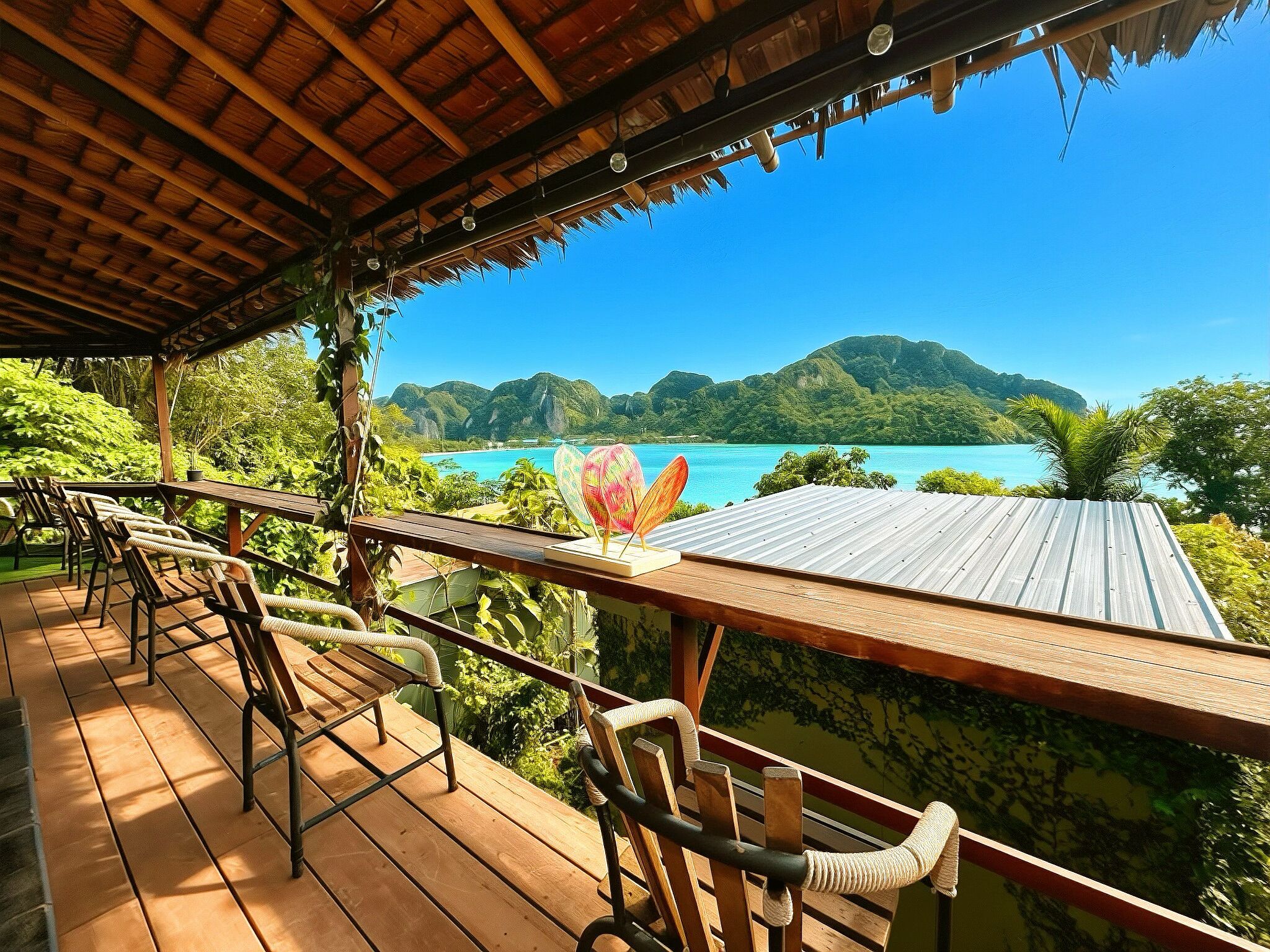 Une terrasse en bois avec des chaises, une table et une vue sur une mer turquoise et des collines verdoyantes.