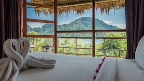 Quarto de hotel com uma janela grande que oferece vista para montanhas exuberantes e um lago.
