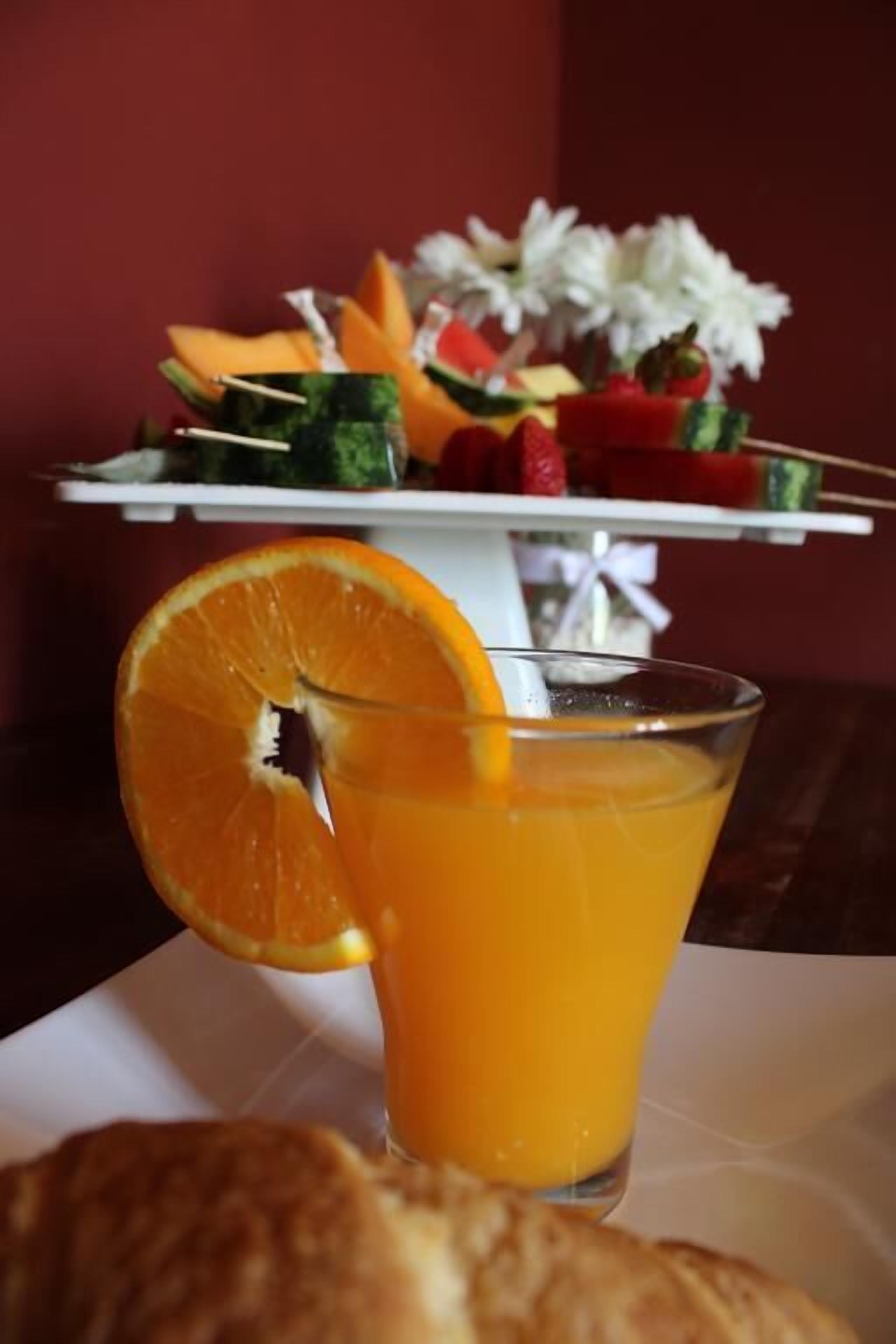Un vaso de jugo de naranja con una rodaja de naranja, un pastel y una fuente de frutas al fondo.