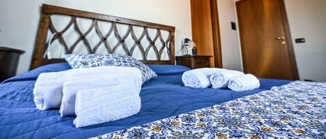 A neatly made bed with blue bedding and white towels, a wooden headboard, and a wooden door in a room with white walls.