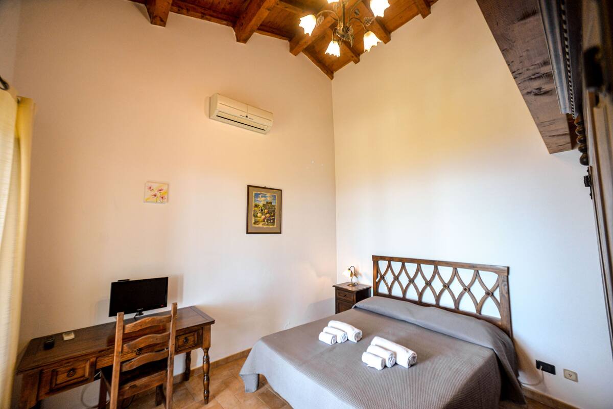 A bedroom with a bed, a desk with a computer, a chair, a nightstand, and a ceiling fan.