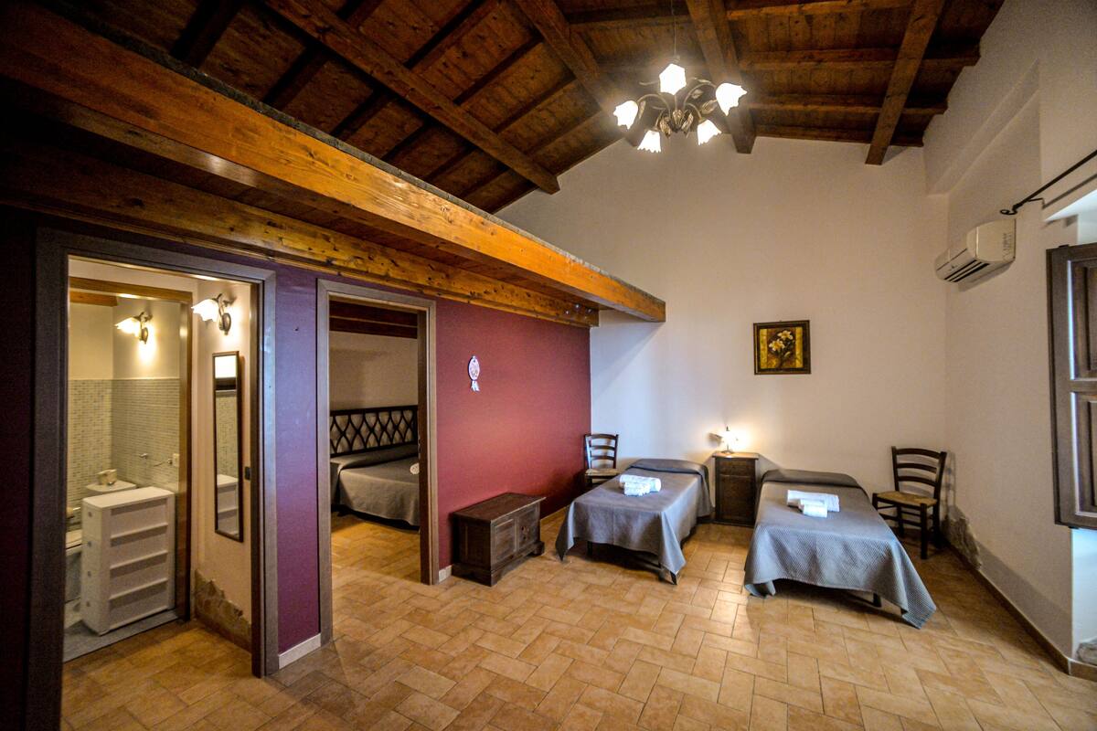 A room with two beds, a small table, a chair, a wooden ceiling, and a bathroom visible through an open door.