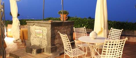 An outdoor patio with a stone fireplace, white metal chairs, and a table.