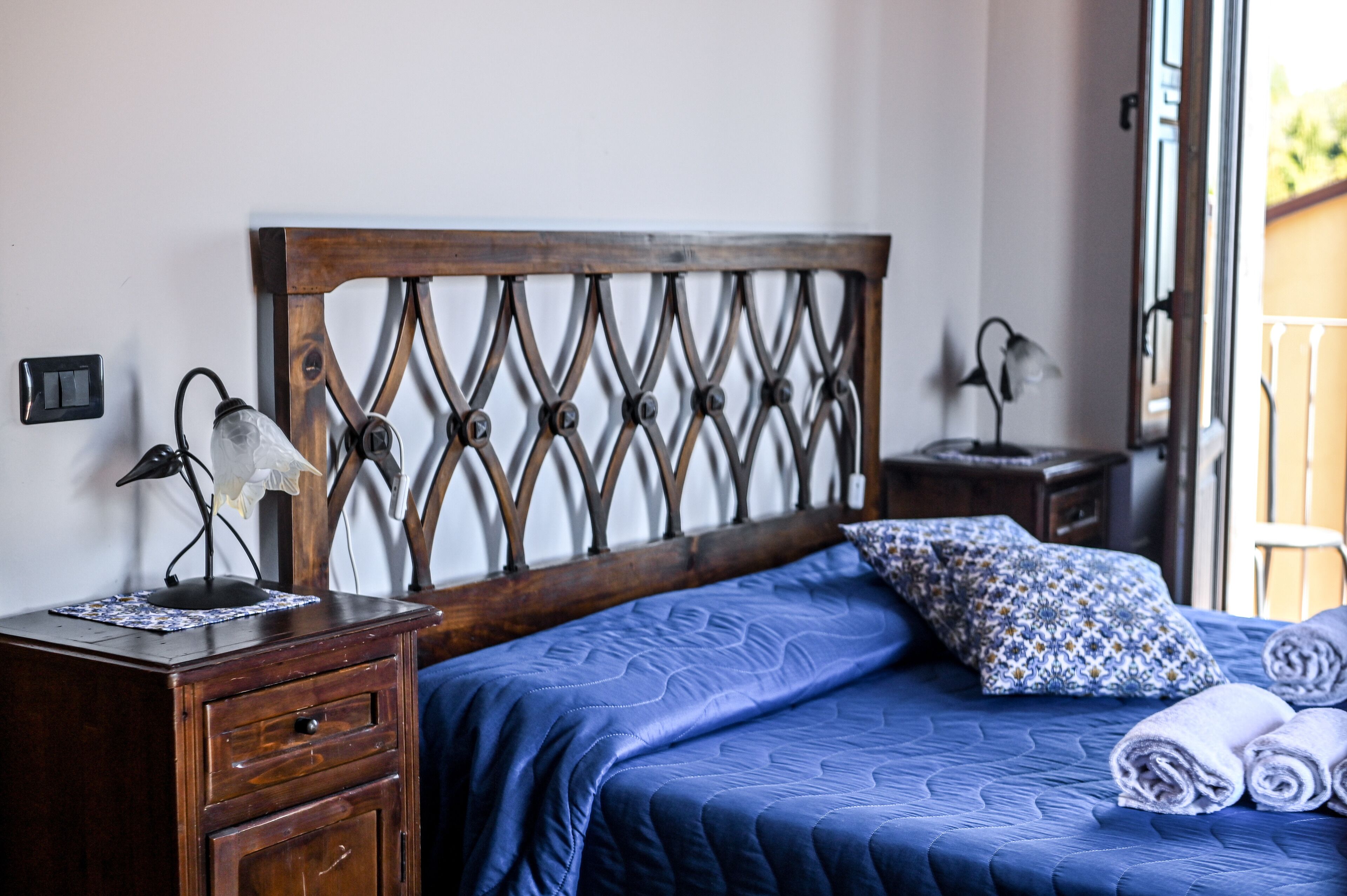 A bedroom with a wooden headboard, a bedside table with a lamp, a patterned pillow, and rolled towels.