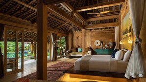Una habitación amplia con una cama grande, vigas de madera, vistas al exterior con vegetación y un comedor con mesa y sillas.