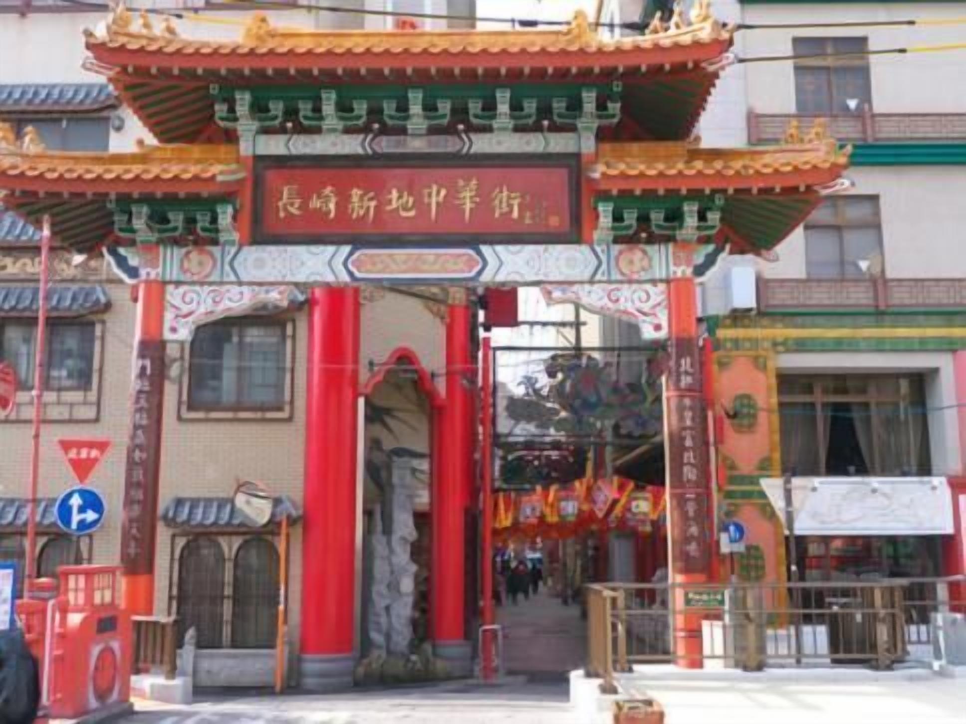 Um portão chinês tradicional, com pilares vermelhos e desenhos detalhados, que dá acesso a uma rua com lojas e uma placa.