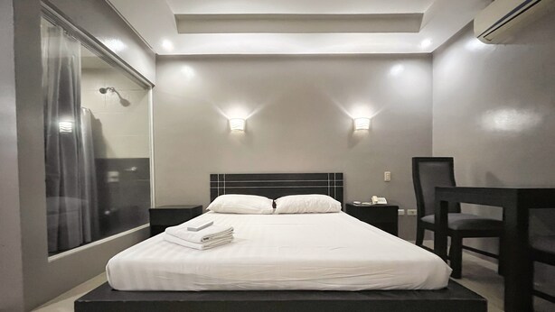 A modern hotel room with a large bed, bedside tables, a desk, and a view through a large window. - Rumi Apartelle Hotel (Angeles City)