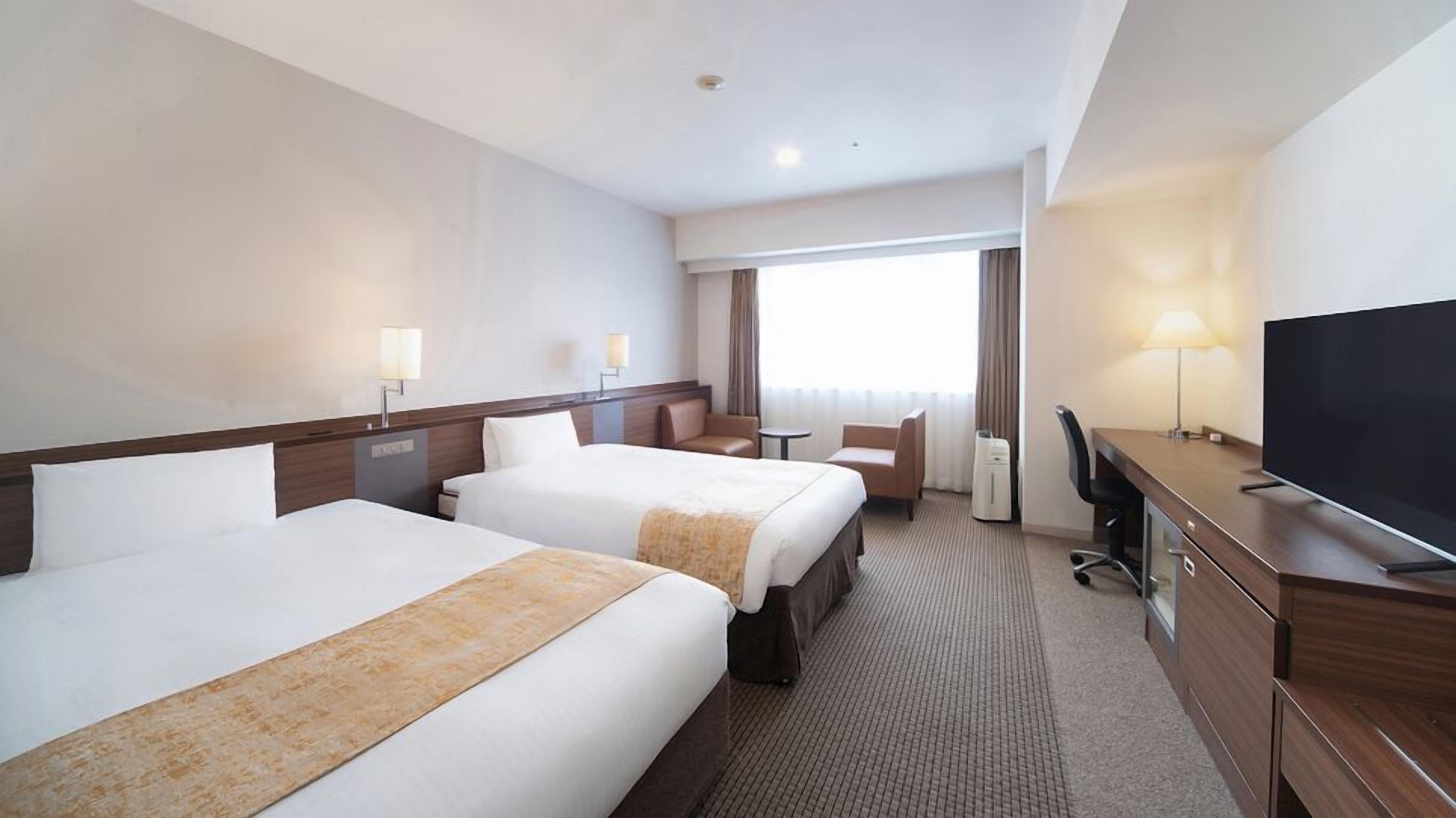 Habitació d'hotel amb dos llits, una taula de treball amb televisió, una cadira, una làmpada i una finestra amb cortines.
