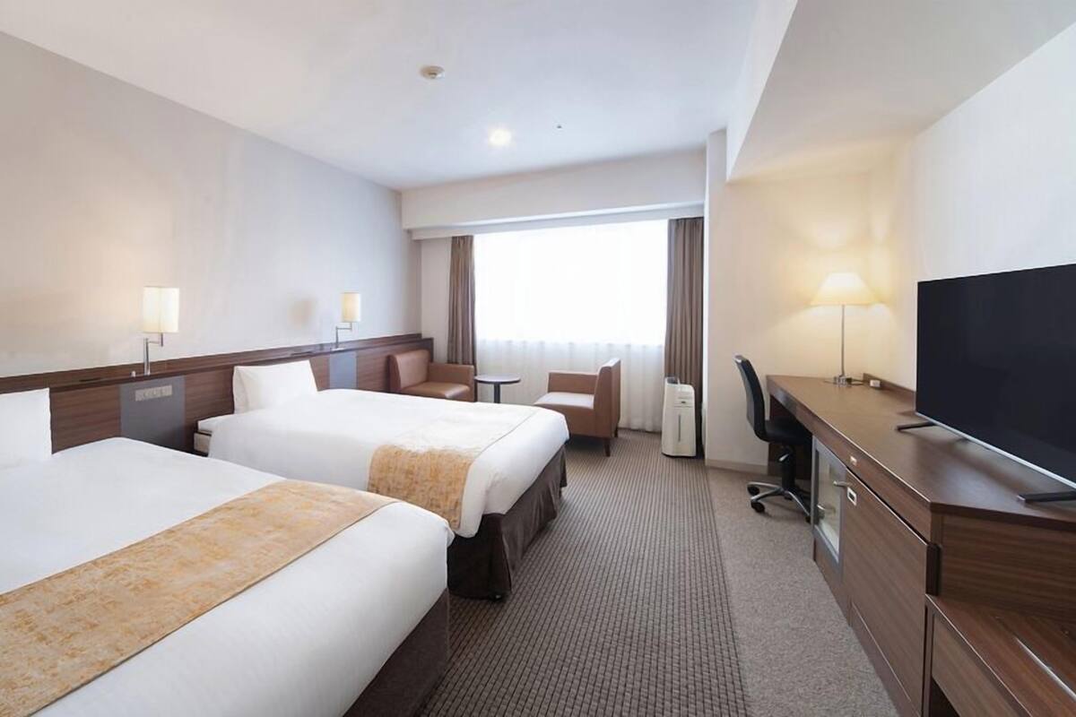 Habitación de hotel con dos camas, un escritorio con televisor, una silla, una lámpara y una ventana con cortinas.