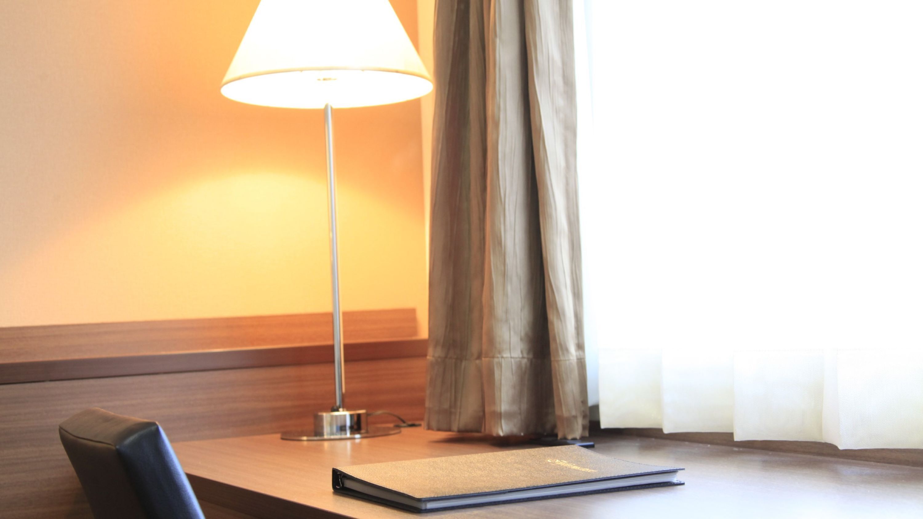 A hotel room with a wooden headboard, a table with a lamp, a notebook, and a curtain.