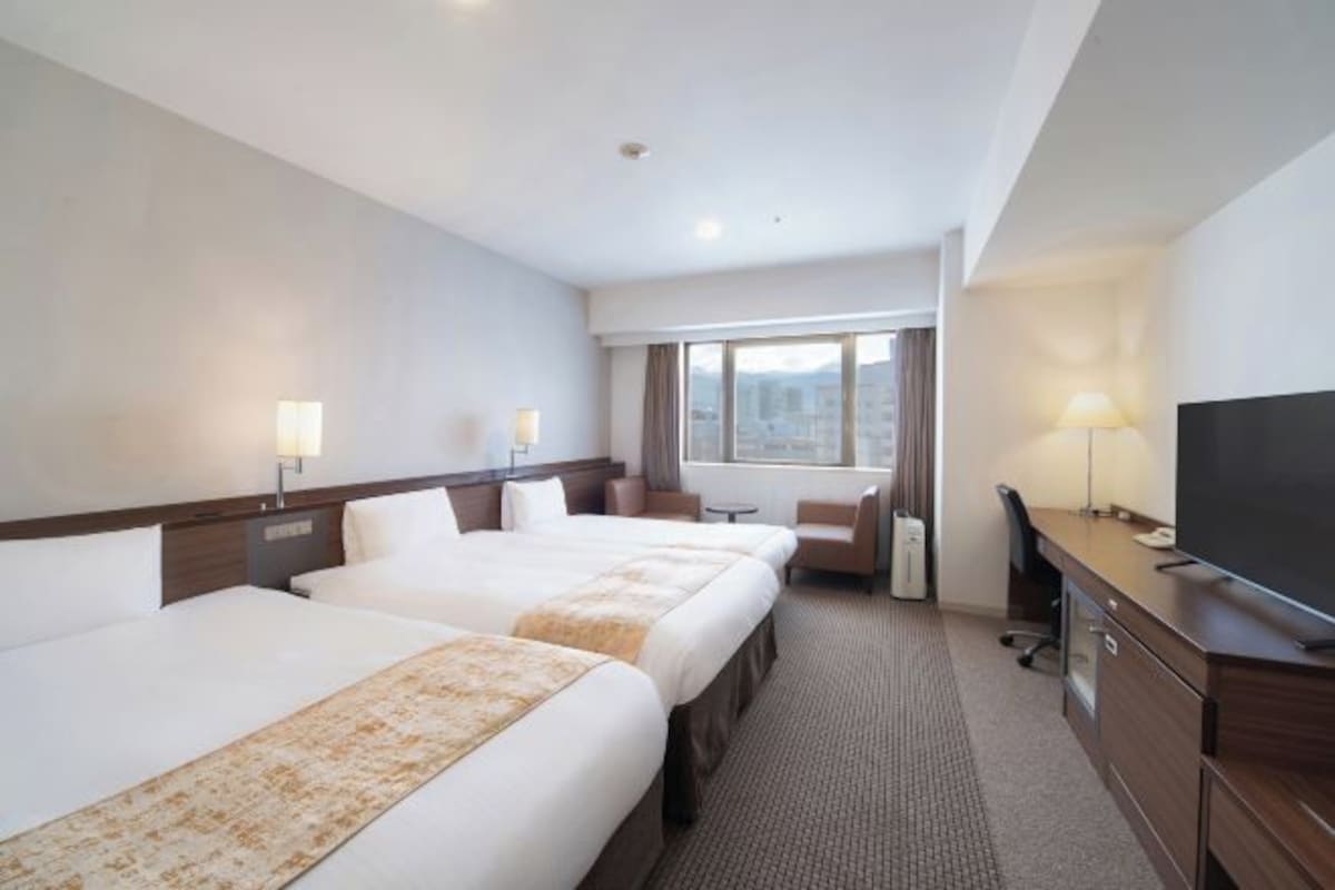 A hotel room with three beds, a desk, a TV, and a window with a city view.