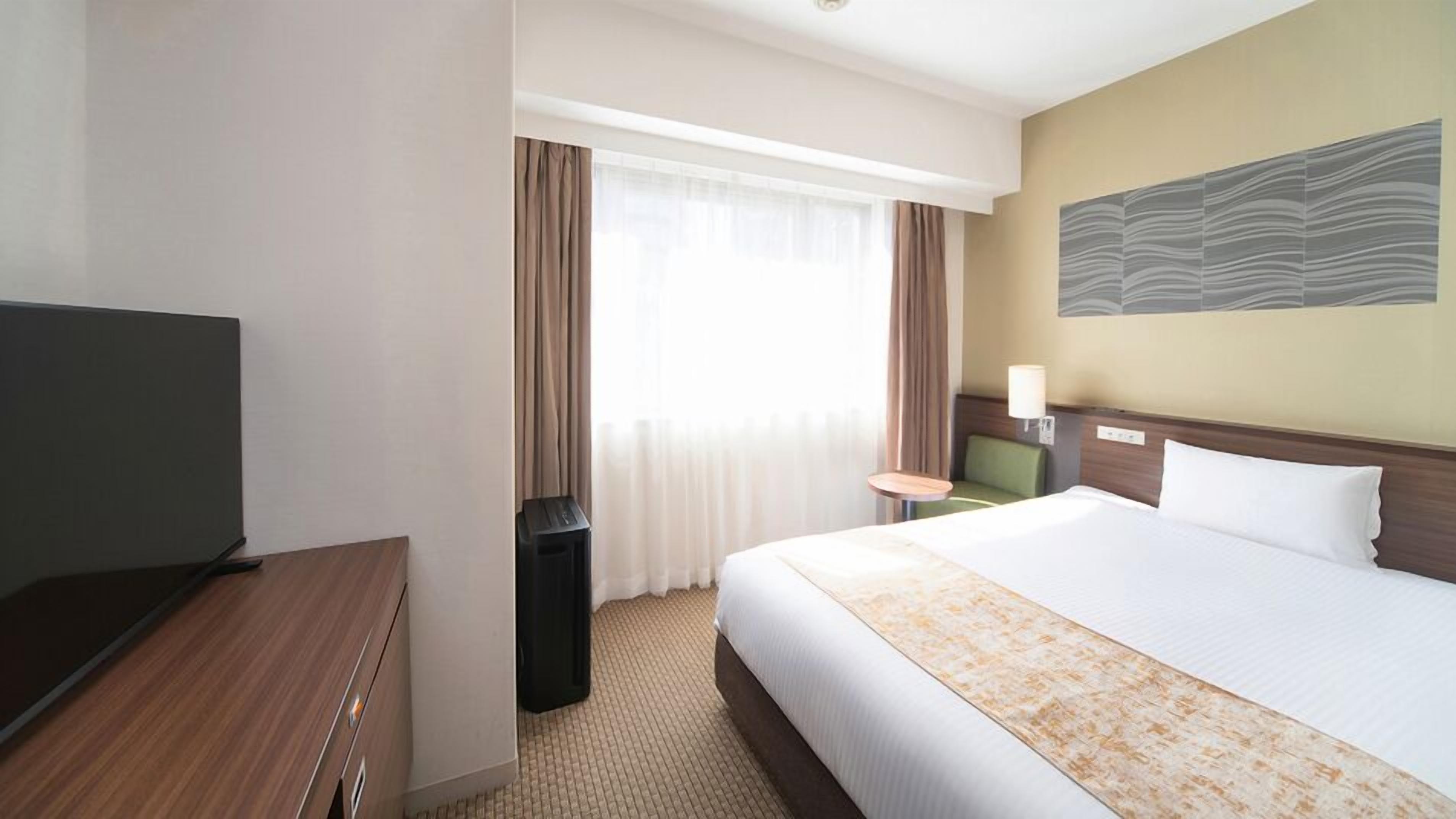 Habitació d'hotel amb llit, escriptori, cadira, televisió i finestra amb cortines.
