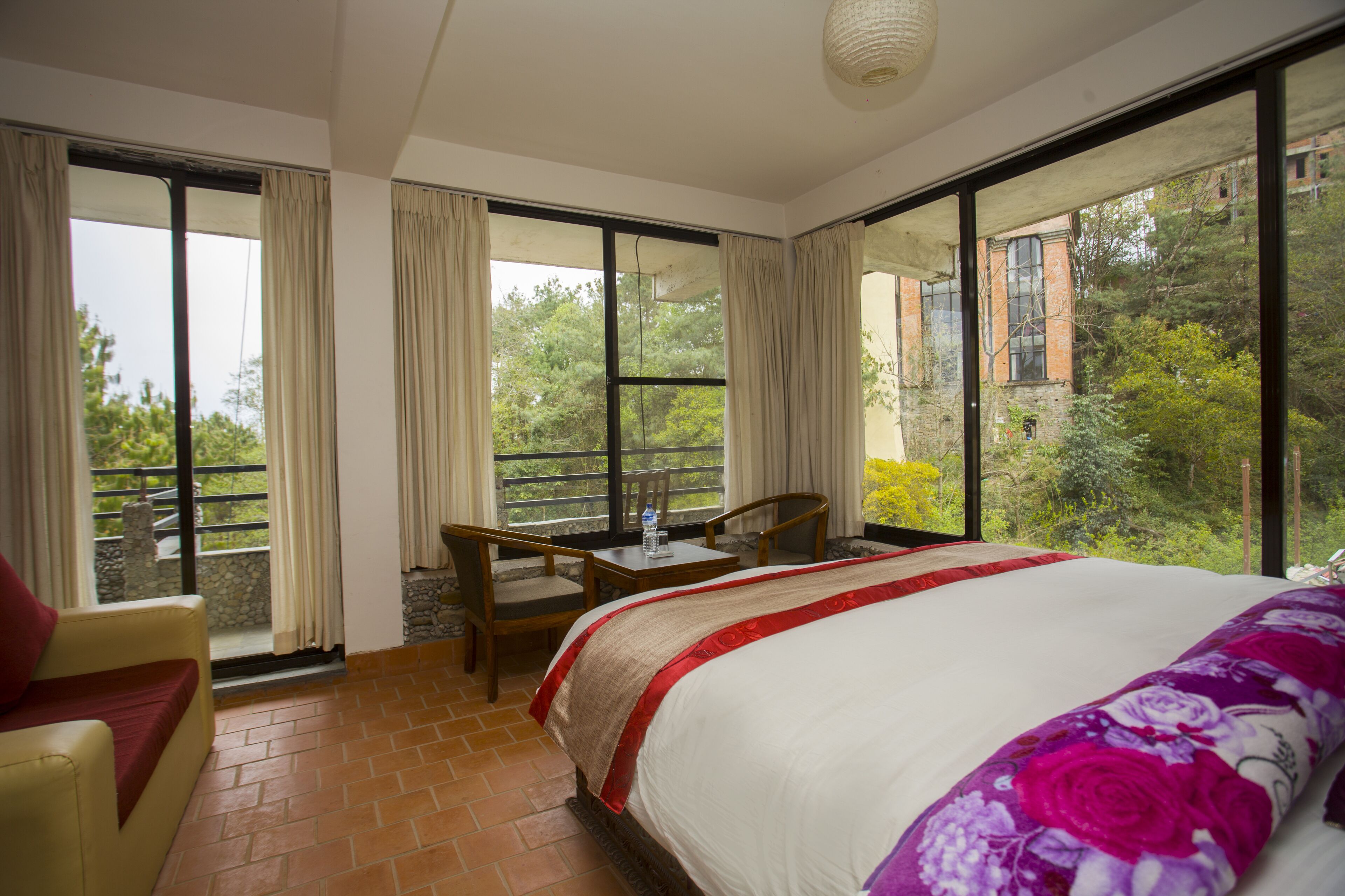A hotel room with a large bed, a chair, a table, and a view of greenery and buildings outside.