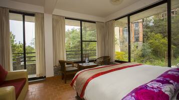 A hotel room with a large bed, a chair, a table, and a view of greenery and buildings outside.