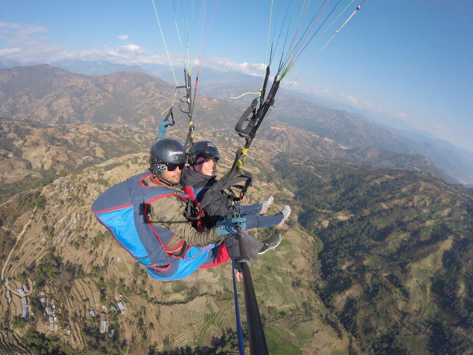 A person paragliding with a harness and parachute, overlooking a mountainous landscape with terraced fields and scattered buildings.