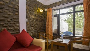 Ein Zimmer mit einer Steinsäule, einem beigen Sofa mit roten Kissen, zwei Holzsesseln, einem kleinen Holztisch und einem Fenster mit Vorhängen.