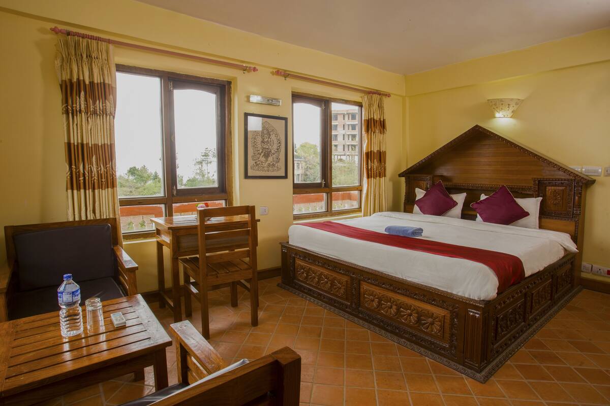Quarto de hotel com uma cama de madeira, uma mesa com cadeira e uma janela com cortinas.