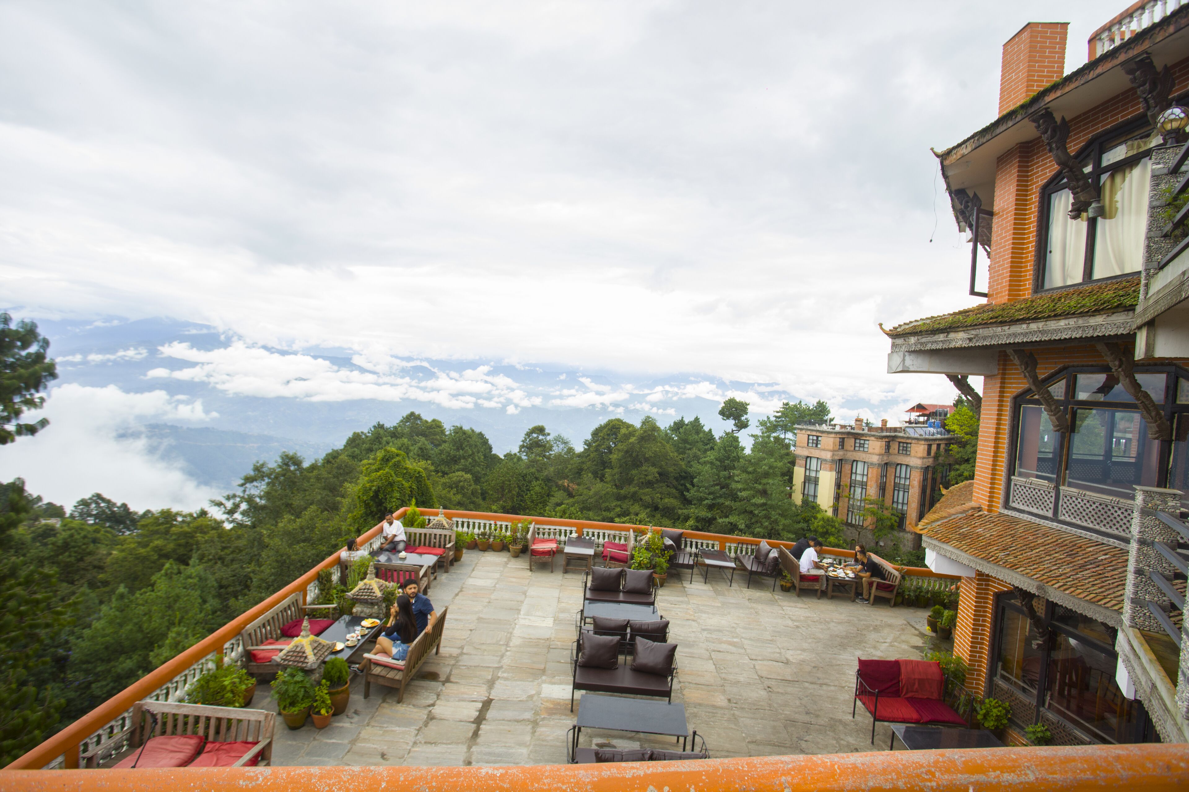 A rooftop terrace with outdoor seating, surrounded by lush greenery and a view of the clouds.