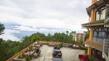 A rooftop terrace with outdoor seating, surrounded by lush greenery and a view of the clouds.