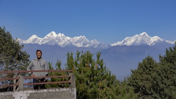 Una persona parada sobre una plataforma de concreto con barandilla metálica, observando un paisaje montañoso con picos nevados.