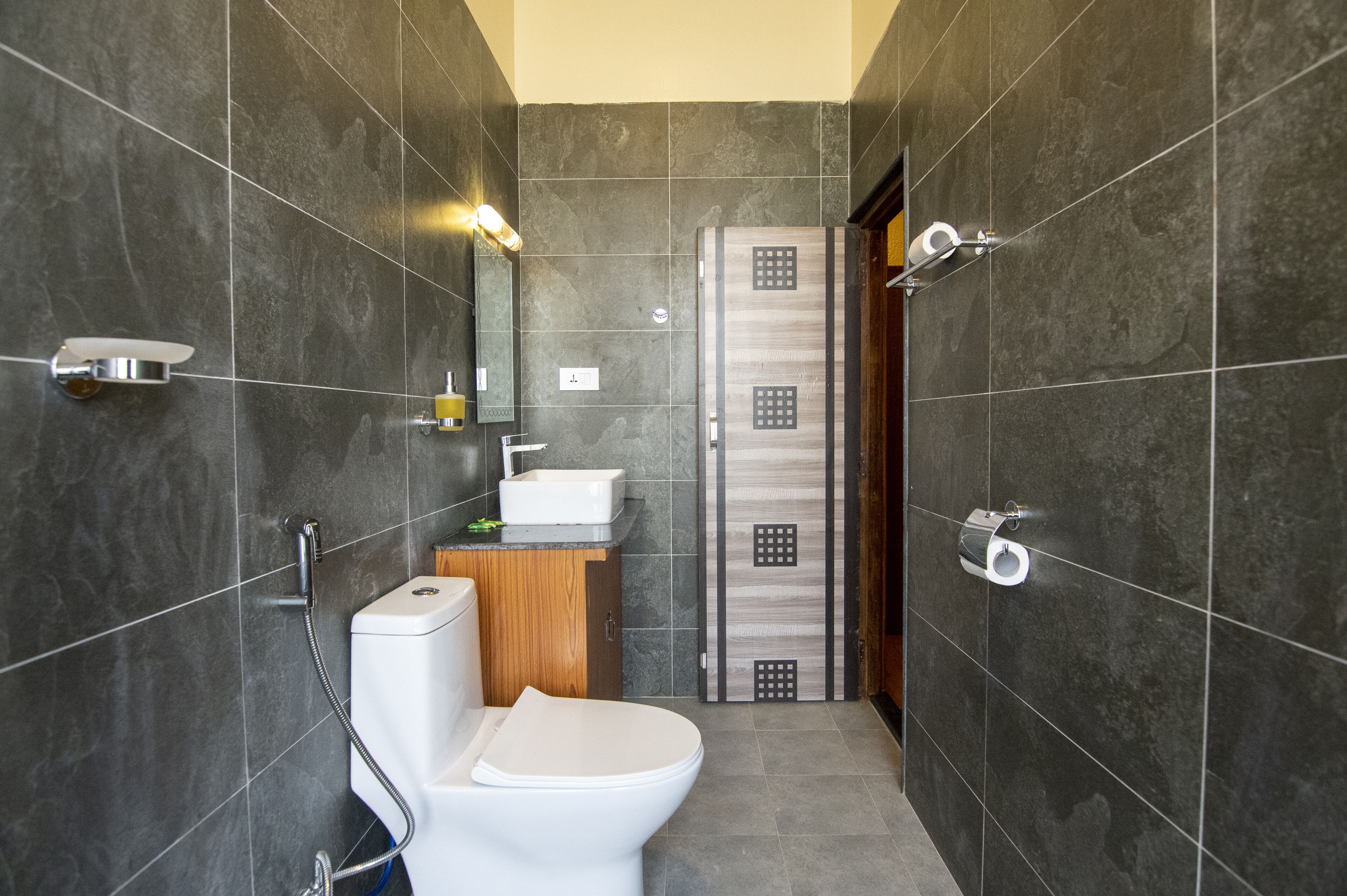 A modern bathroom with dark tiles, a white toilet, a wooden cabinet, and a shower area.