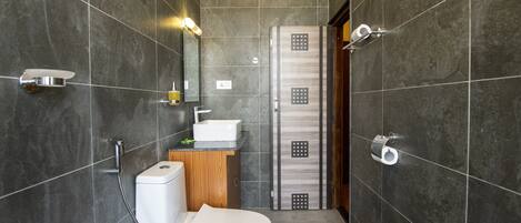 A modern bathroom with dark tiles, a white toilet, a wooden cabinet, and a shower area.