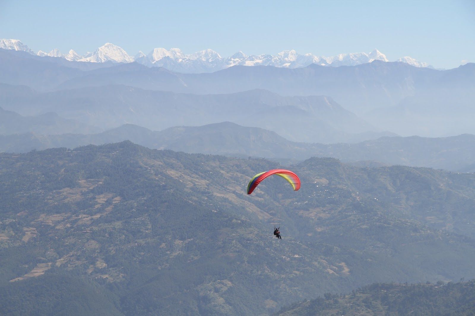 Eine Person gleitet mit einem Paragleiter über eine gebirgige Landschaft, im Hintergrund schneebedeckte Gipfel.
