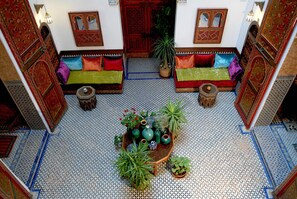 Un interior tradicional marroquí con suelo de azulejos, una zona de estar central con cojines y varias plantas en macetas.