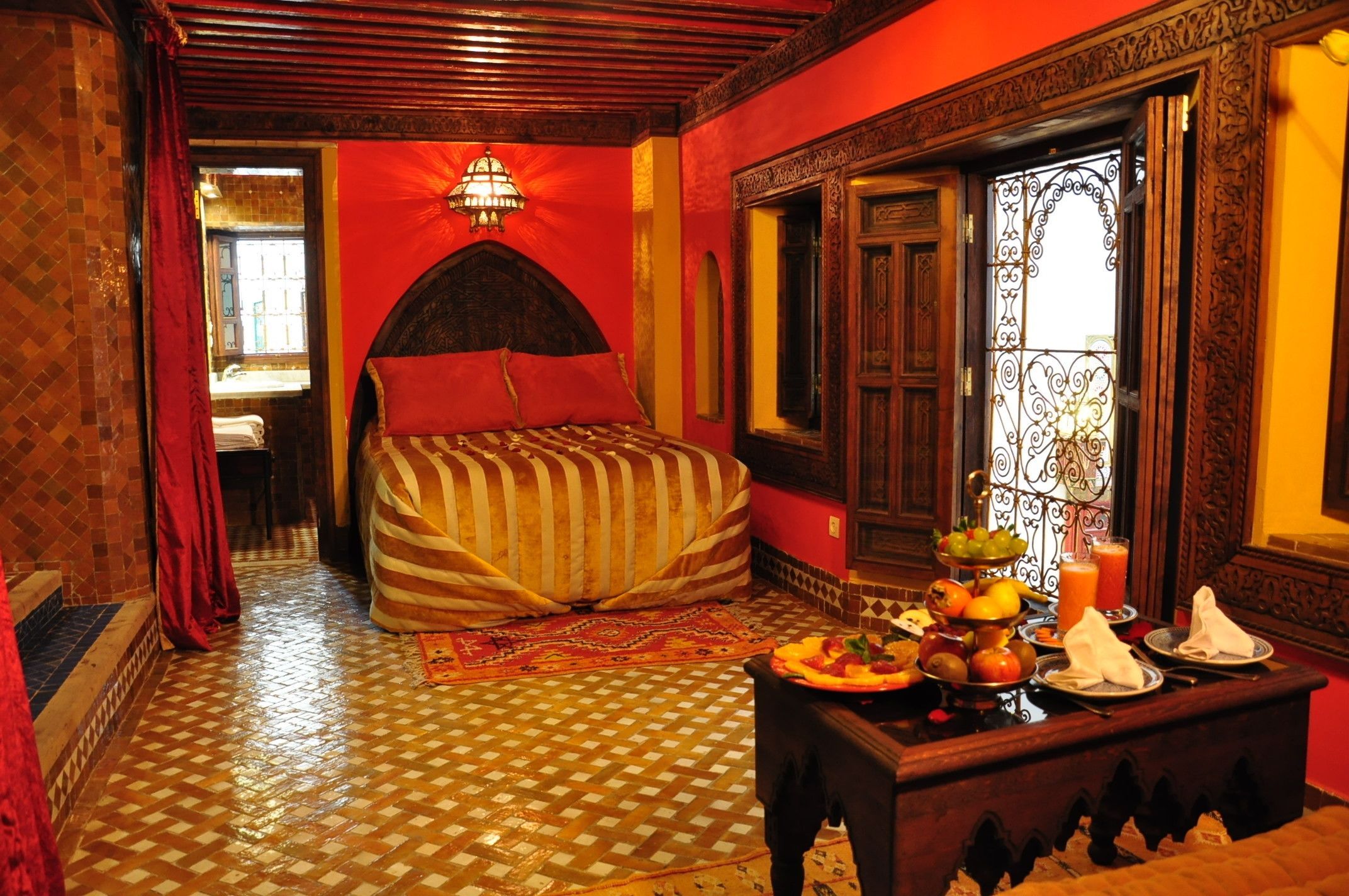 A room with a checkered floor, a bed with striped bedding, a table with fruits and drinks, and a decorative mirror.