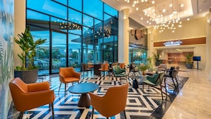 Een moderne hotellobby met grote ramen, een vloer met een geometrisch patroon en diverse zithoekjes, waaronder ronde tafels en stoelen.