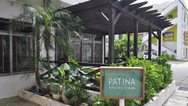 Ein Gebäude mit überdachter Außenbereiches, einem Schild mit der Aufschrift "PATINA" und verschiedenen Pflanzen.