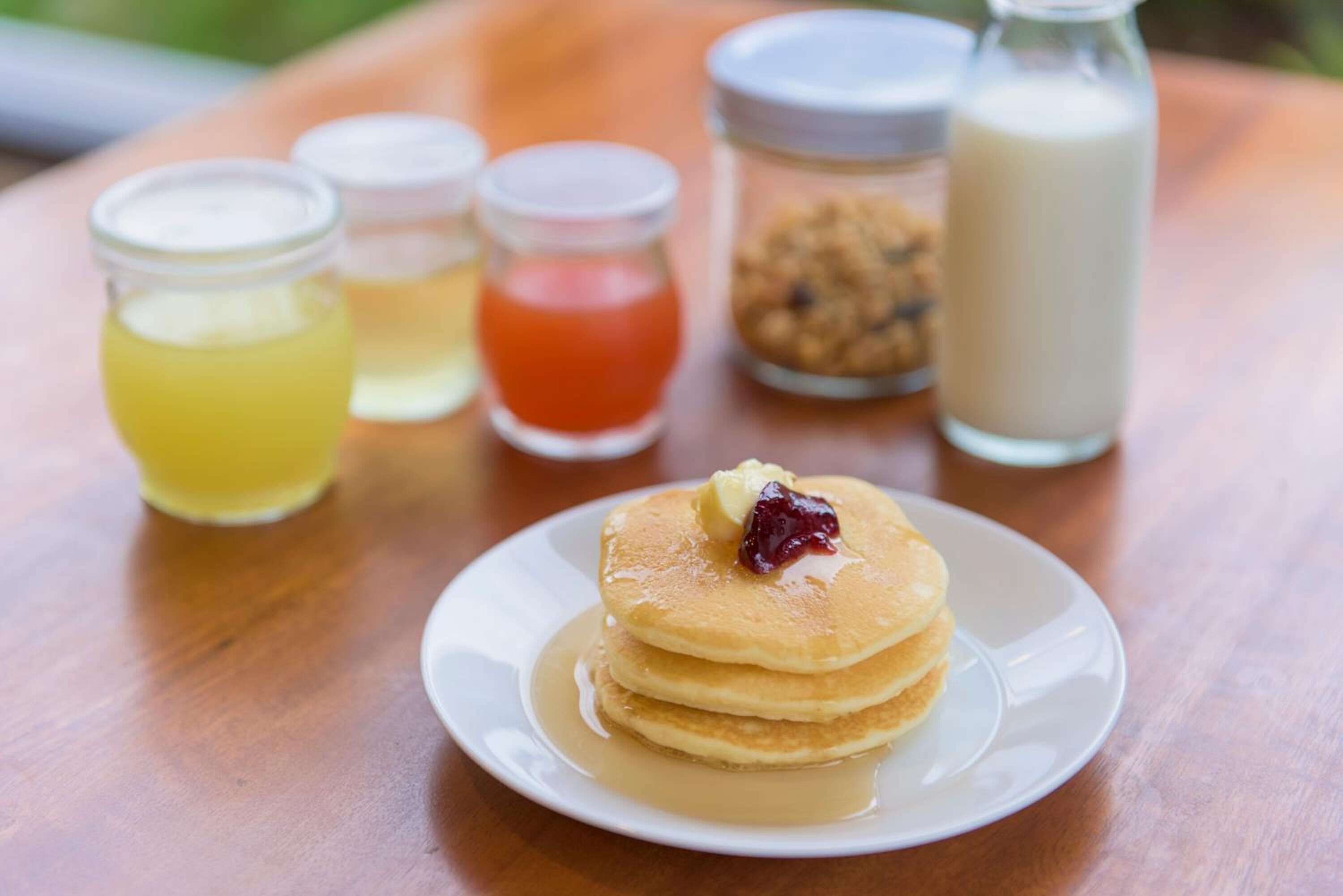 Une pile de crêpes sur une assiette blanche, nappée de sirop et surmontée d’une noisette de beurre, accompagnée de trois petits pots de condiments et d’une bouteille de lait, le tout posé sur une table en bois.