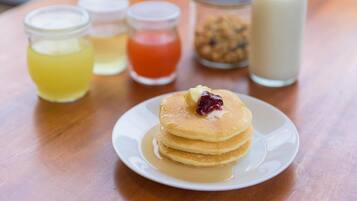 Une pile de crêpes sur une assiette blanche, nappée de sirop et surmontée d’une noisette de beurre, accompagnée de trois petits pots de condiments et d’une bouteille de lait, le tout posé sur une table en bois.