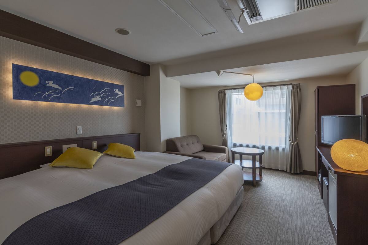 A hotel room with a large bed, a small round table, a sofa, a TV, and a window with curtains.