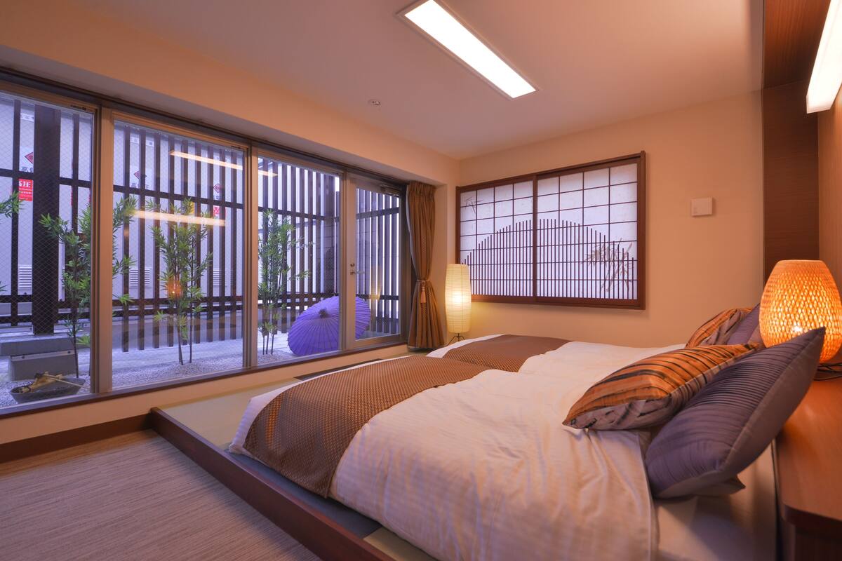 A hotel room with a large window, a bed with multiple pillows, a nightstand with a lamp, and a view of a fence and plants outside.