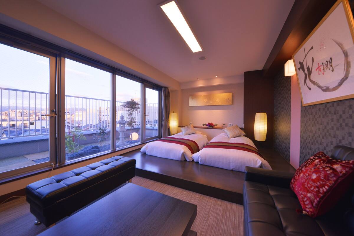 A modern hotel room with a large window, a sofa, a bed with white linens, and a red pillow.