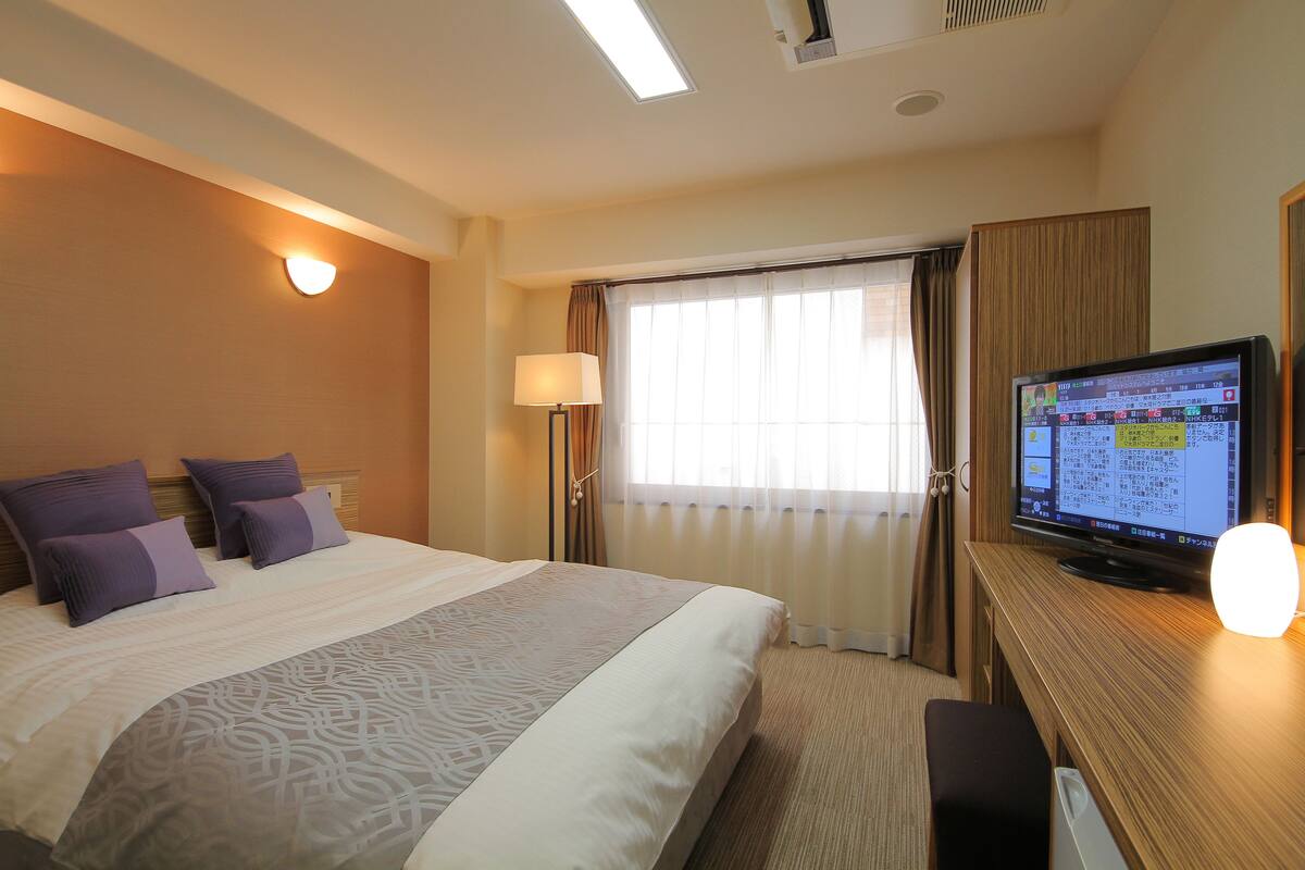 A hotel room with a bed, a desk with a computer, a lamp, and a window with curtains.