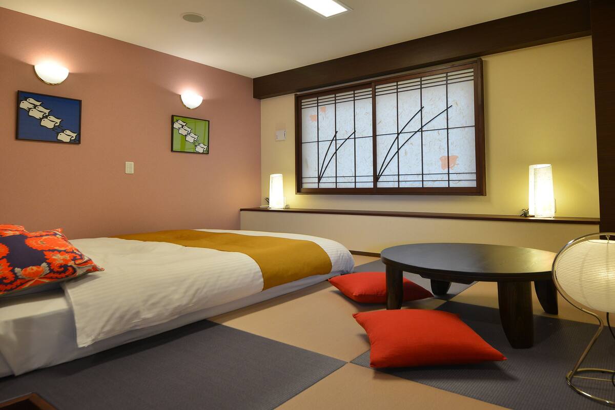 A hotel room with a bed, a small table, and a chair. There are two wall sconces providing light.