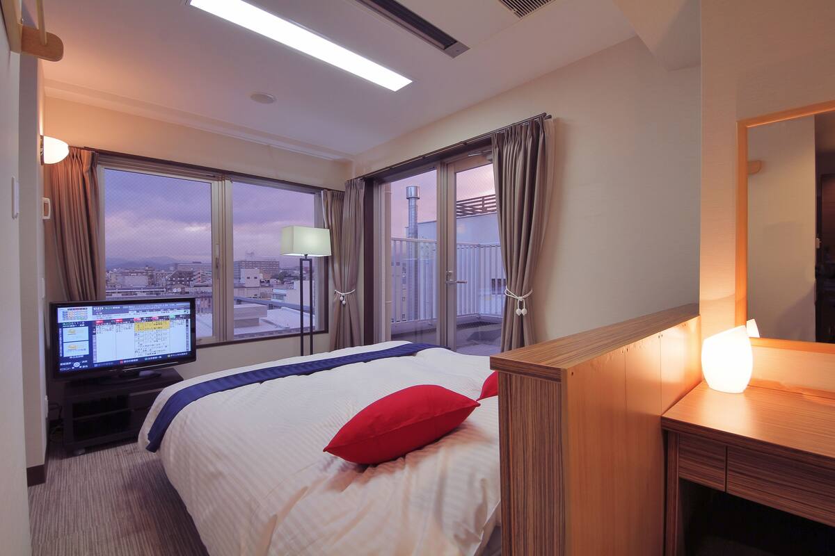 A hotel room with a bed, a desk, a television, and large windows offering a city view.