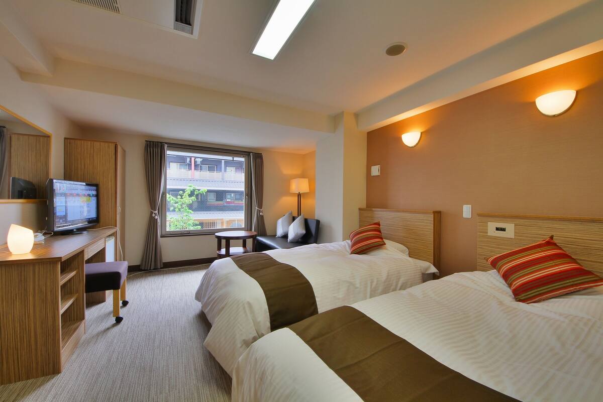 A hotel room with two beds, a desk with a television, a chair, and a window with curtains.