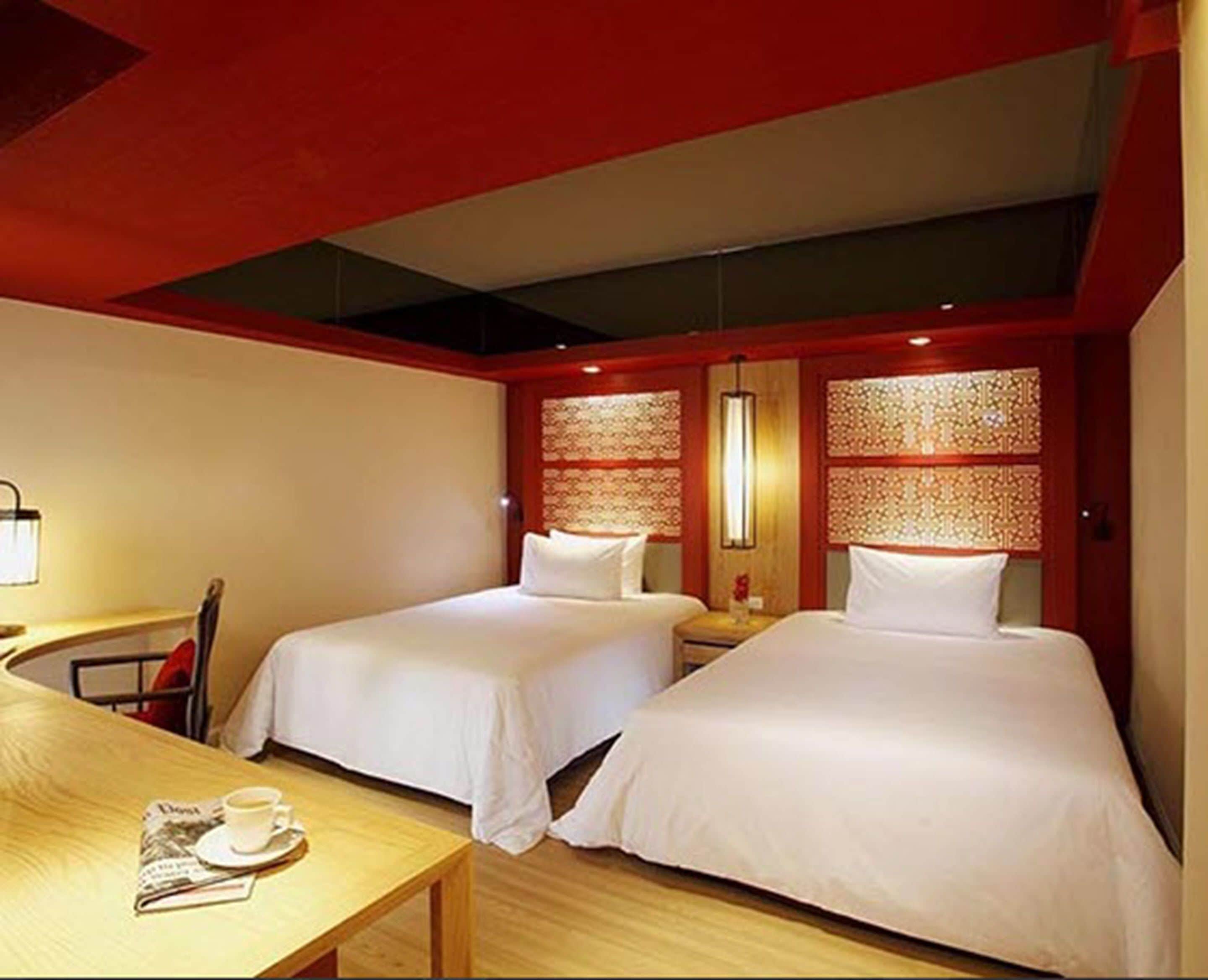 A hotel room with two beds, a wooden table with a newspaper and a cup, and a red accent wall.