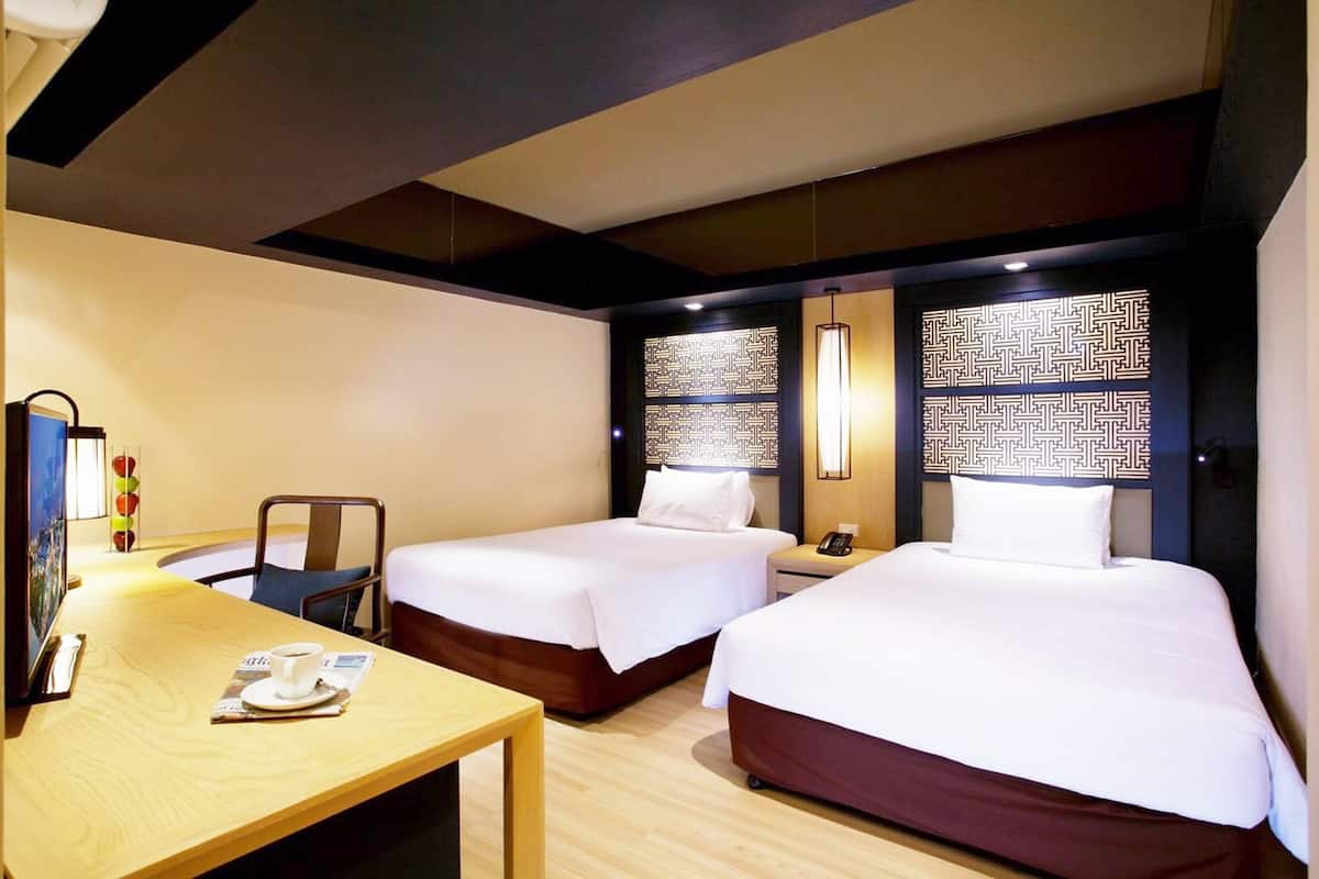 A hotel room with two single beds, a wooden desk, a chair, and a small table with a cup and a magazine.
